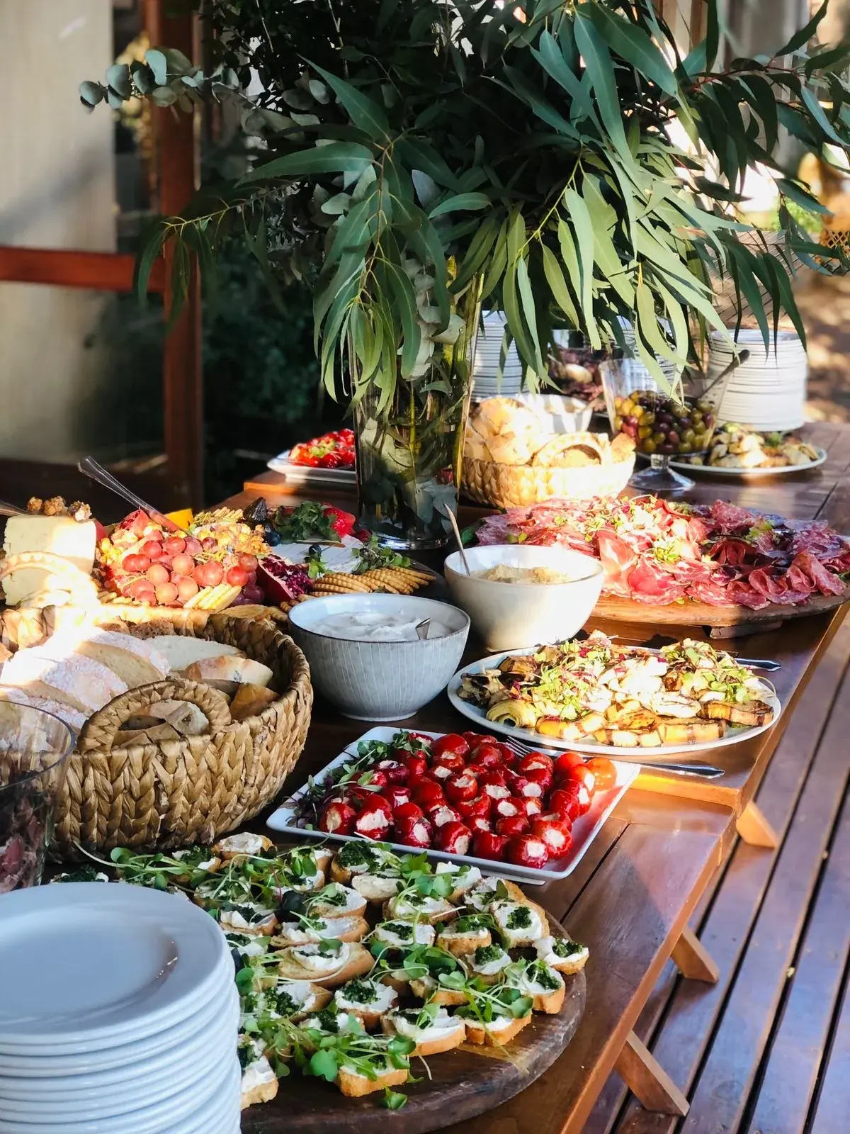 A wooden harvest table with bread baskets, cheeses, sliced meats, dips, red peppers and topped bread bites