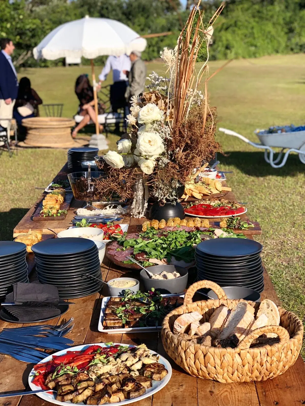 An outdoor food table on a lawn with bread baskets, platters, stacked black plates and white roses