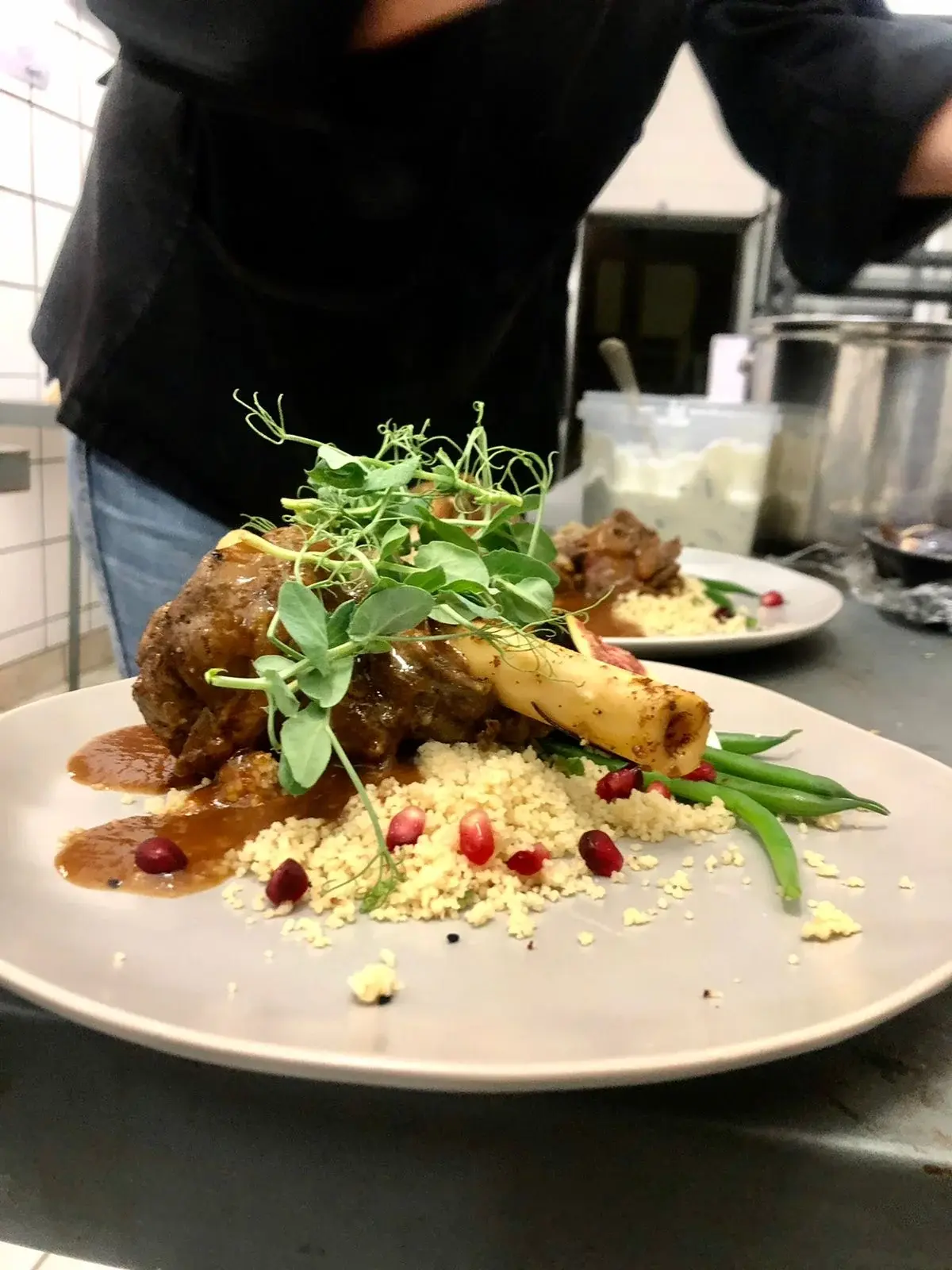 A plated meat shank with couscous, green beans, pomegranate seeds and microgreens on a kitchen counter