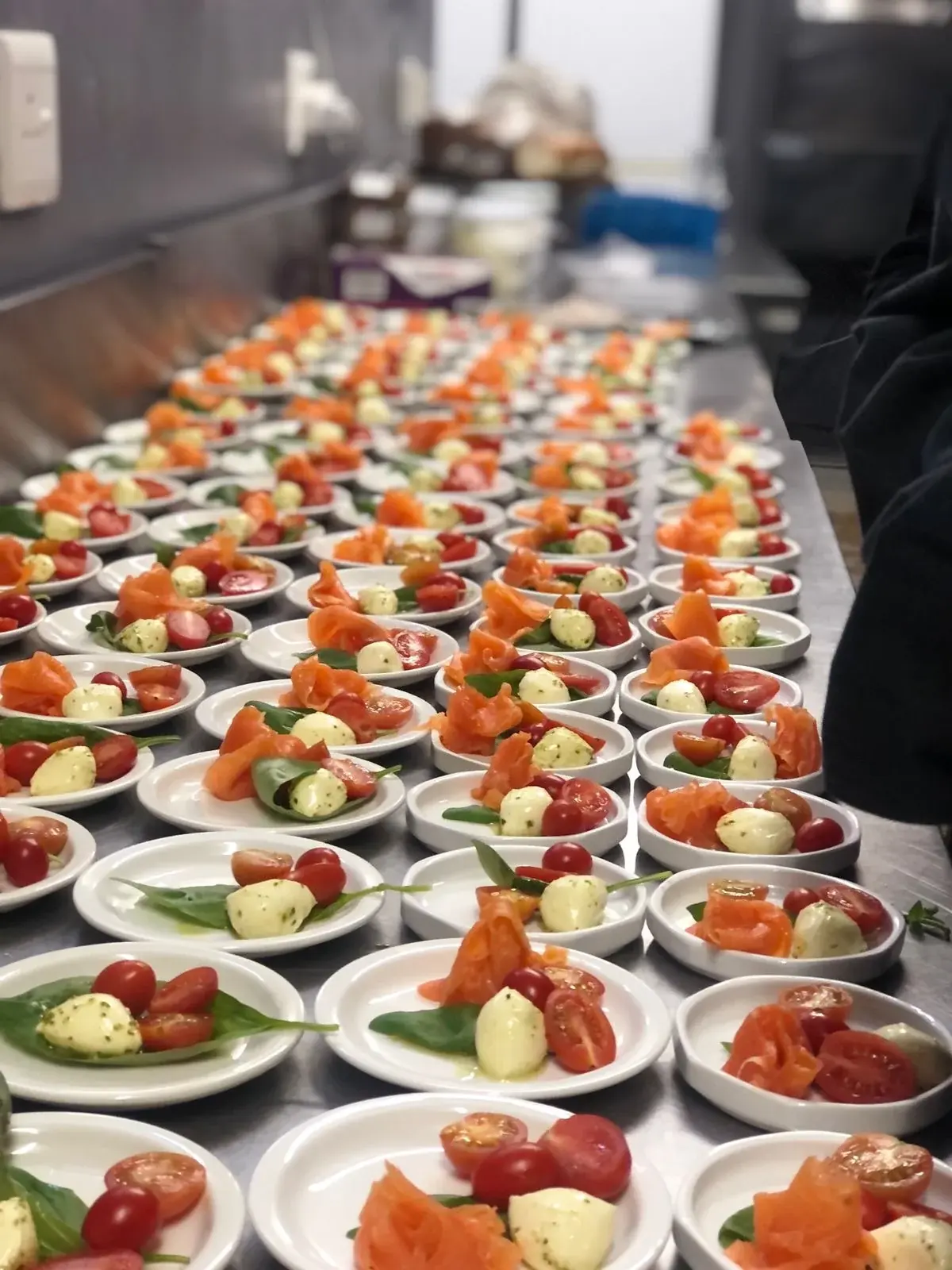 Rows of small white plates with salmon-coloured slices, cherry tomatoes, pale round bites and green leaves